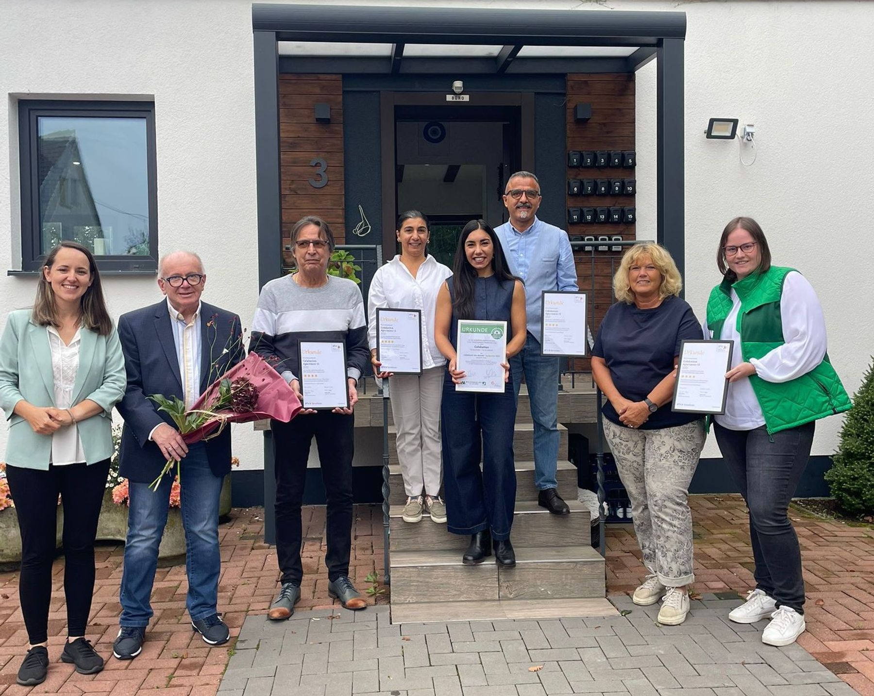Eight people stand in front of a building, some holding certificates and one holding a bouquet of flowers. They are smiling and posing for a group photo on a paved entrance.